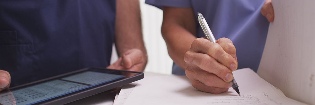 Close-up shot of hands of health workers in scrubs - one holds file and pen, other holds tablet